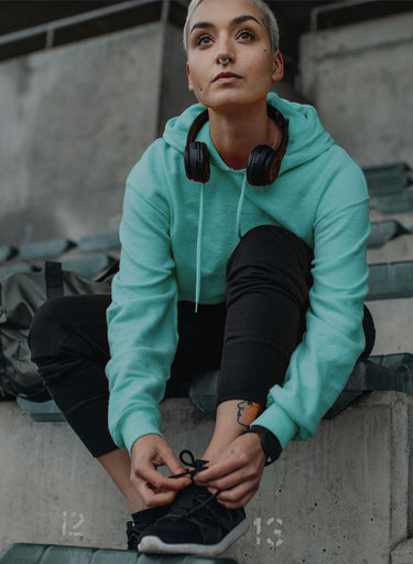 A woman on stadium bleachers fastening the laces of her KSNS shoes.