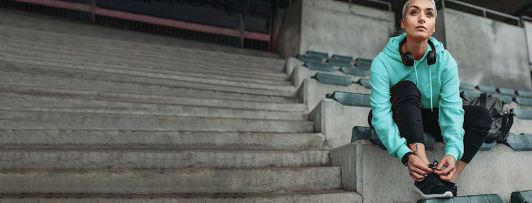 A woman on stadium bleachers fastening the laces of her KSNS shoes.