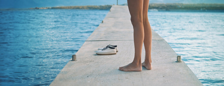 A woman stands barefoot on a dock, with KSNS shoes placed nearby.